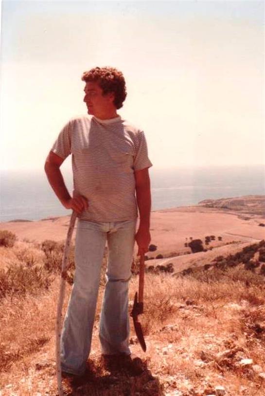 A young adult on a hillside gazes at the ocean horizon, casually holding tools.