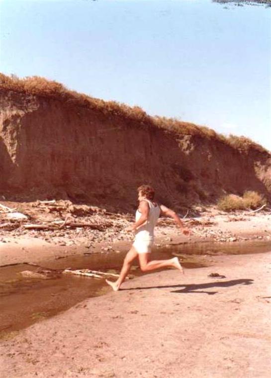 Individual joyfully running by a stream on a sunny day near sandy cliffs.
