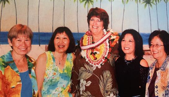 Three women stand smiling together in festive clothing near a beach-themed backdrop.