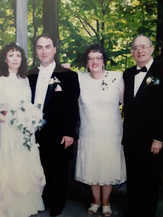 Couple in wedding attire stands with two guests in formal wear surrounded by greenery.