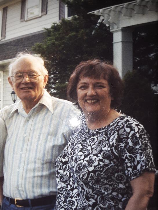 Two elderly individuals stand closely together, smiling warmly in a garden setting.