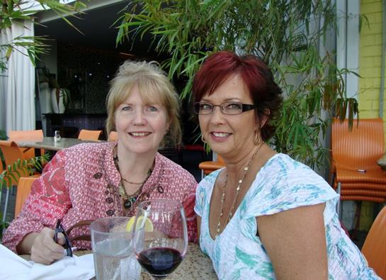 Two happy women enjoy wine at a lively outdoor cafe amidst lush greenery.