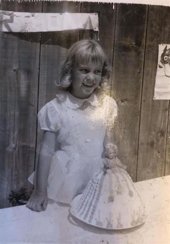 Young girl smiles while holding her doll, dressed in a white outfit, with a wooden wall behind.