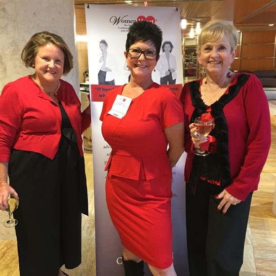 Three women dressed in elegant red outfits gather for a celebration, holding glasses and smiling.