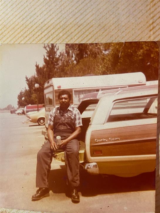 A man relaxes on a parked car trunk, enjoying a sunny day in a bustling parking area.