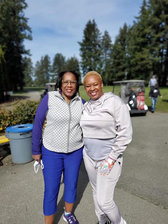 Two friends smile in casual clothes amidst a lush green landscape.