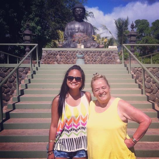 The women stand together near a towering statue surrounded by lush greenery and blue skies.