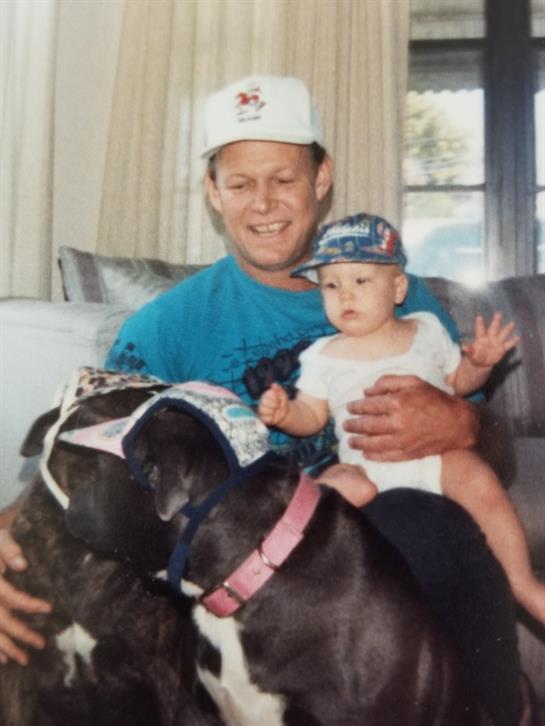 A joyful man holds a baby while two friendly dogs gather around them in a warm setting.