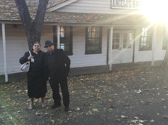 A couple stands smiling by an old building as leaves fall around them in the warm glow of sunset.