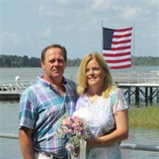 A couple enjoys their wedding anniversary by a peaceful lake, proudly standing with a bouquet.