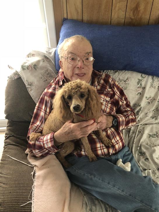 An elderly man sits comfortably on a couch, holding a poodle with a gentle smile during the day.