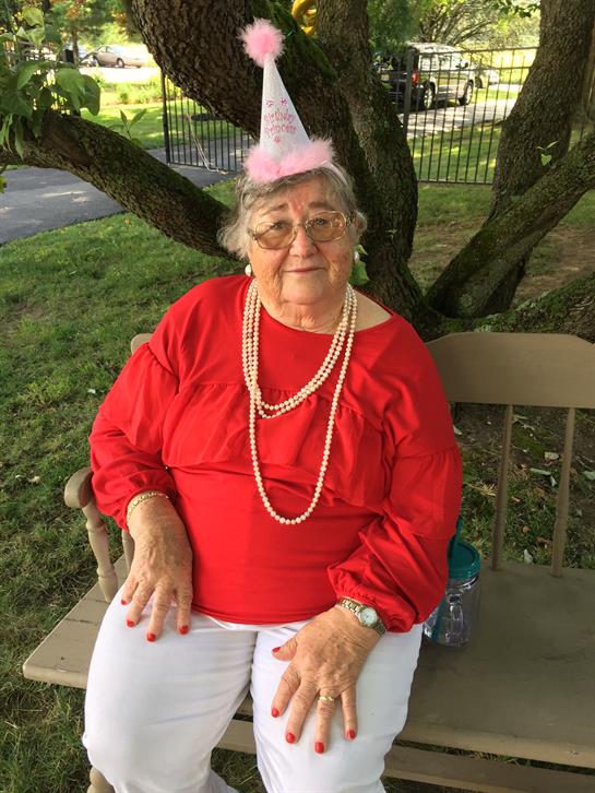 An elderly woman in a red dress and pearls enjoys a joyful moment in a park, wearing a festive hat.