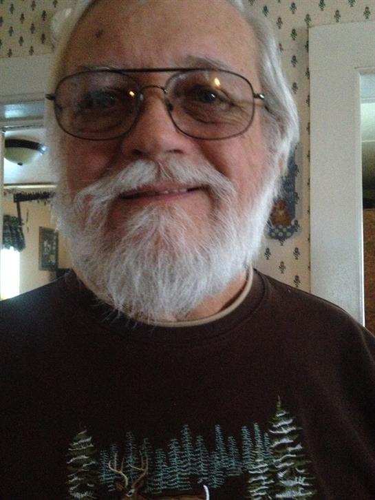 An elderly man with a white beard and glasses smiles in a bright, floral-patterned room.