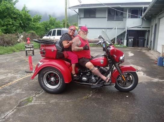Two individuals dressed in red sit on a vibrant motorcycle trike in a tropical setting.