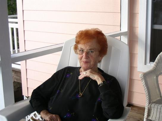 An elderly woman with orange hair sits calmly on a porch chair, lost in thought.