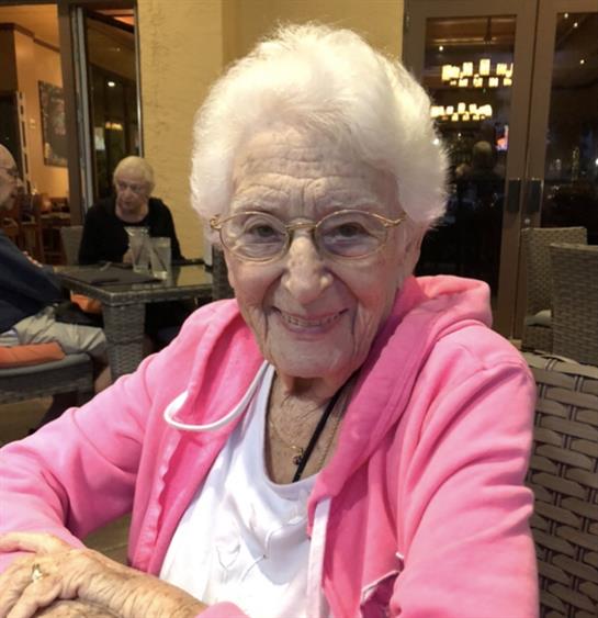 Elderly woman wearing a pink sweater smiles warmly while sitting at a cafe with friends.