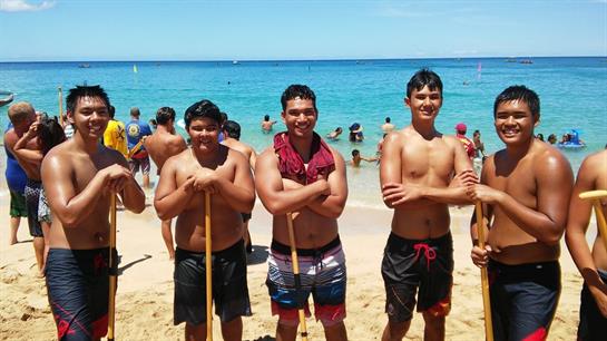 Five young men on a sandy beach hold paddles and smile as others swim nearby.