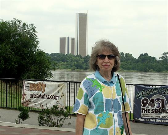 A senior woman in sunglasses stands by a riverbank with a city skyline and greenery.
