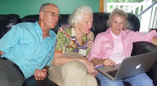 Three older adults are gathered on a couch, discussing something on a laptop, showing interest.