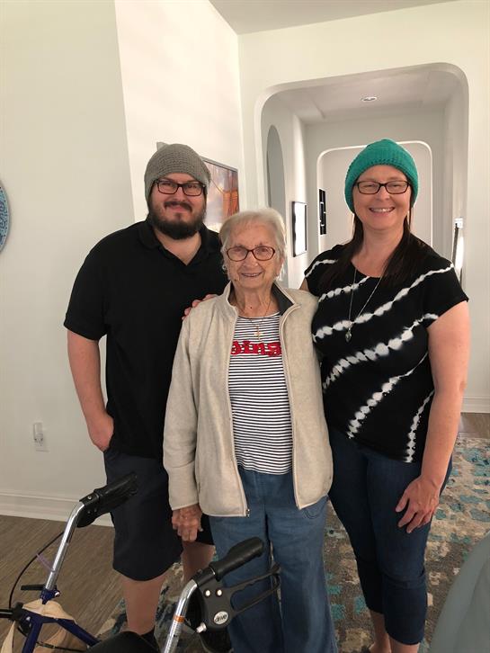 A young man, woman, and elderly lady pose together in a bright, welcoming living room.