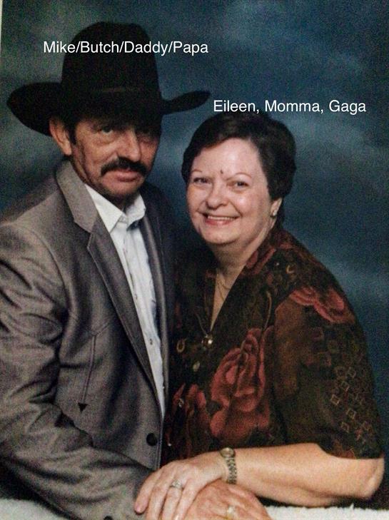 A man in formal attire and a woman in a floral blouse smile while posing closely together.