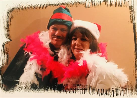 A couple wears festive hats and feather boas, smiling joyfully during a holiday event.