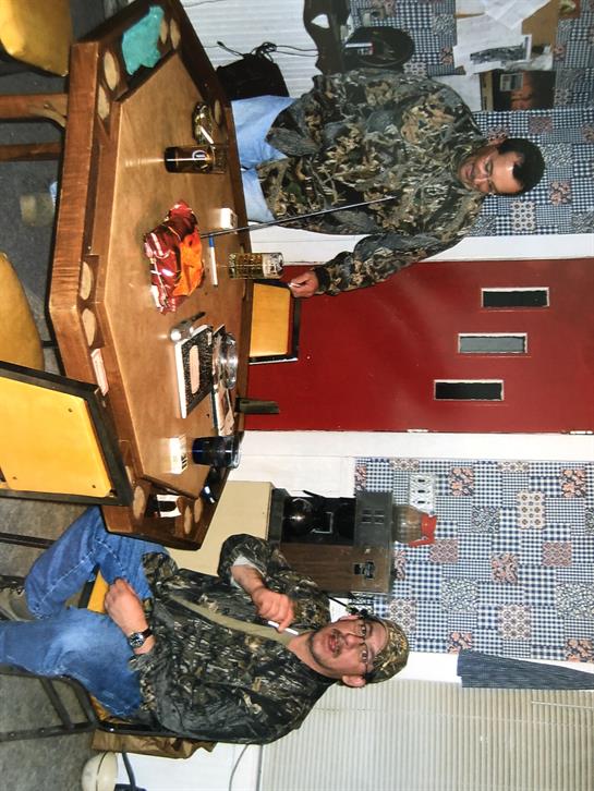 Two men in camouflage clothing relax in a rustic dining area with food and drinks on the table.