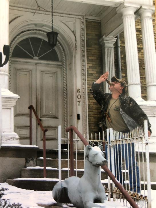 A man dressed in camouflage salutes while standing on the steps of a historic house during winter.