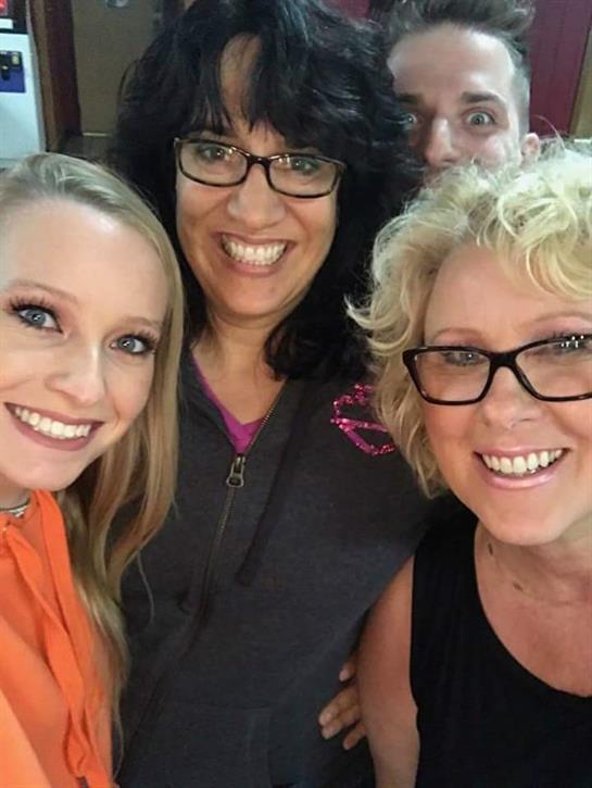 Three women pose happily for a selfie while a fourth person humorously peeks behind them.