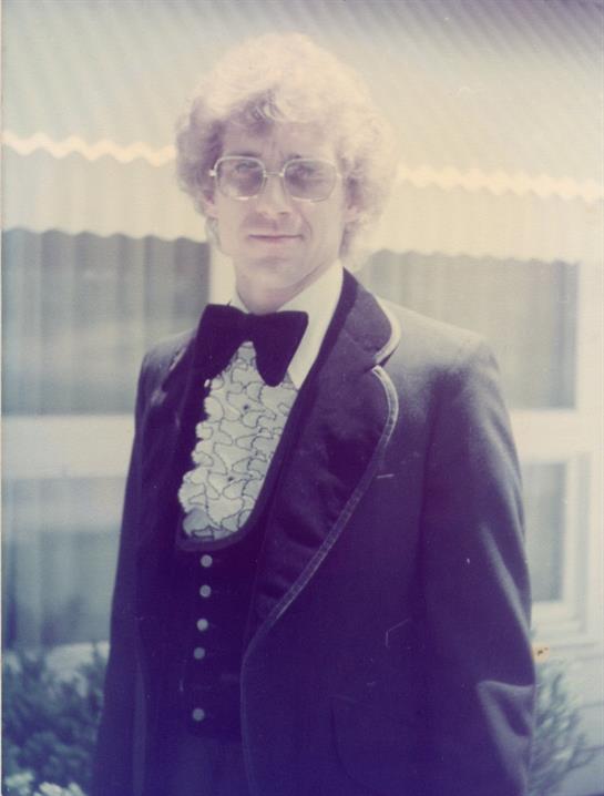 A man confidently wears a ruffled shirt in a formal outfit outdoors in the sun.