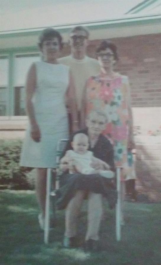 Four women stand around an older woman holding a baby in a stroller, smiling in a green backyard.