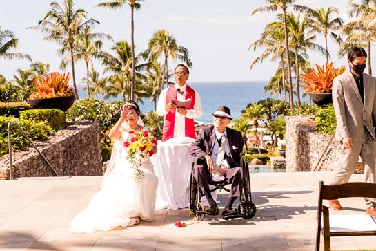 A couple gets married on a sunny day with palms swaying and ocean waves in the background.