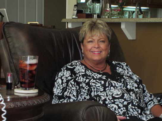 A woman is smiling and seated in a comfortable chair with a drink beside her, enjoying time indoors.