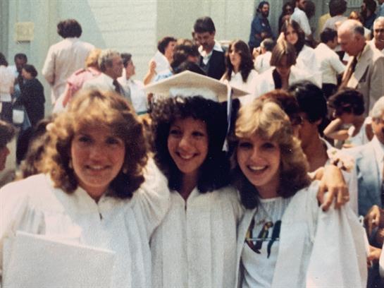 Three friends in white gowns celebrate their achievement outdoors with smiles and laughter.