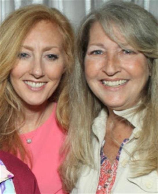 Two women pose happily while enjoying a social moment at a gathering in a sunlit setting.