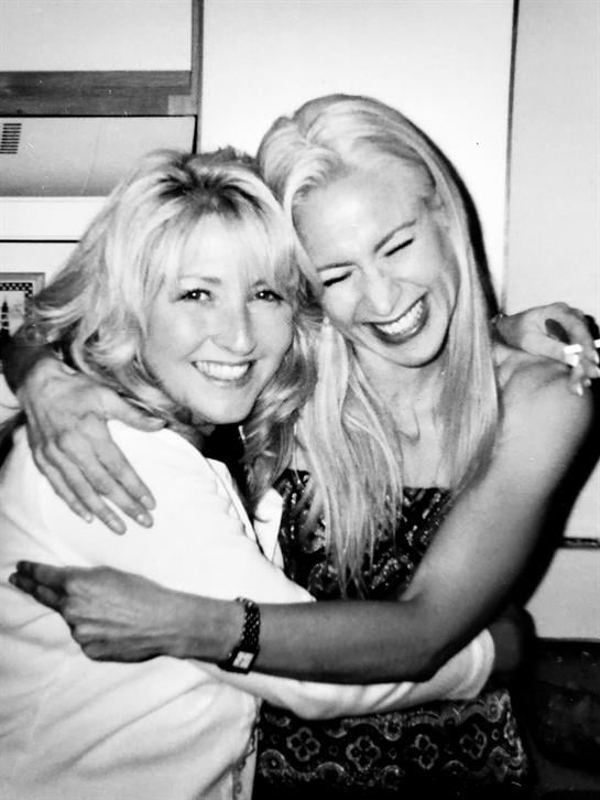 Two women smile and embrace, showcasing their friendship and joy during a lively gathering.