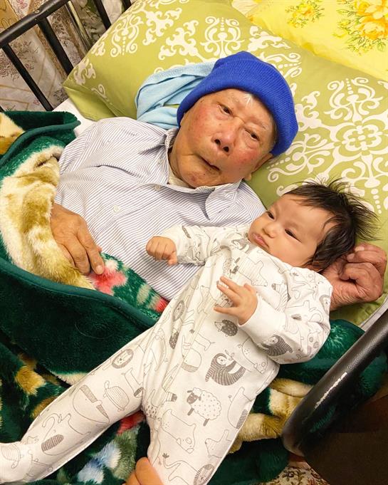 An elderly man relaxes with a baby beside him under a warm blanket in winter.