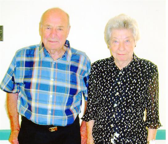 An elderly couple stands side by side, smiling, dressed in casual clothing, and looking cheerful.