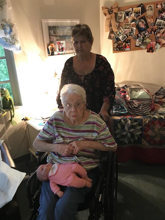 An elderly woman in a wheelchair gently holds a baby doll, with her caregiver nearby.