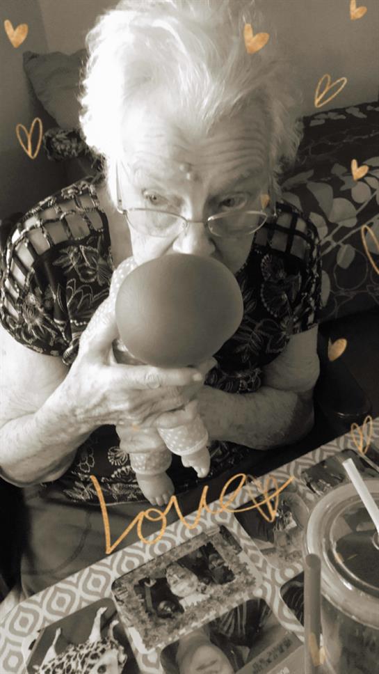 An elderly woman smiles while playfully interacting with a doll at a family gathering.