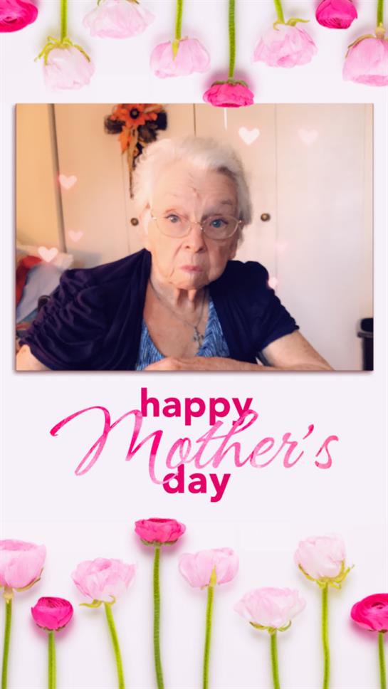An elderly woman sits at a table, warmly celebrating Mother's Day with decorations in a cozy room.