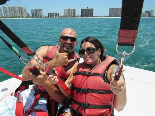 Two people are smiling and posing on a boat, ready for a parasailing experience on sunny day.