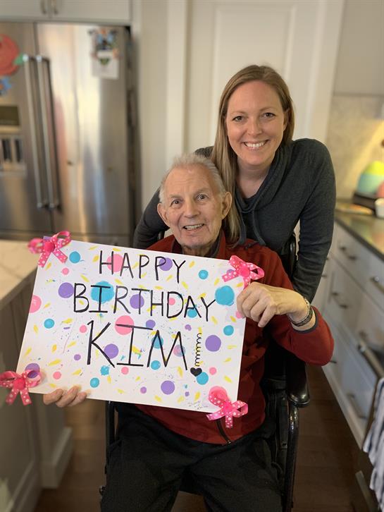 A cheerful woman holds a colorful birthday sign with an elderly person in a kitchen.