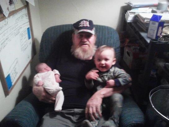A grandfather smiles while holding a baby and sitting next to a toddler in a relaxed atmosphere.