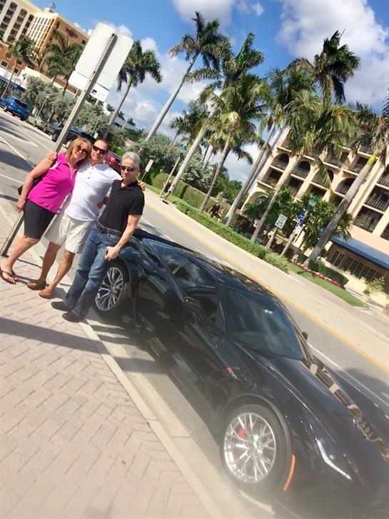 Three friends stand on a sidewalk beside a sleek black car, enjoying a sunny day in a vibrant area.
