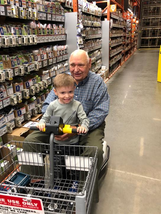 Elderly man and young boy smile while riding a mobility scooter down an aisle filled with supplies.