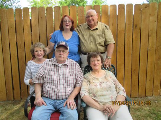 Six family members pose together, smiling happily in a backyard setting surrounded by greenery.