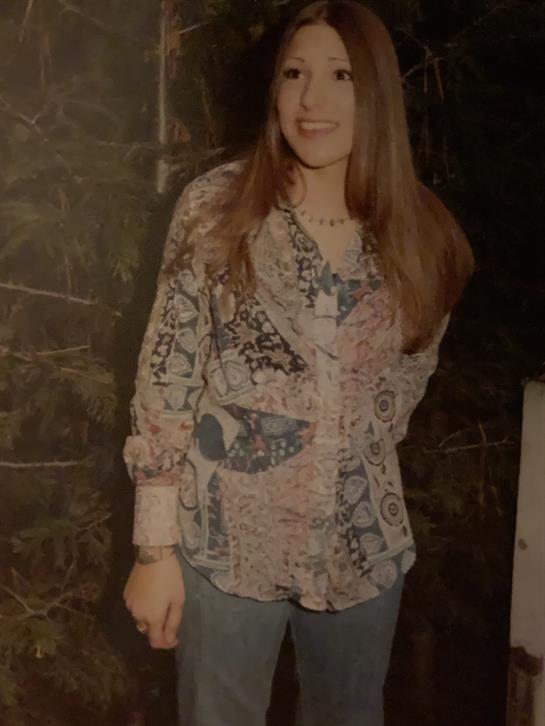 A woman stands confidently in a vintage patterned shirt, smiling in a dimly lit environment.
