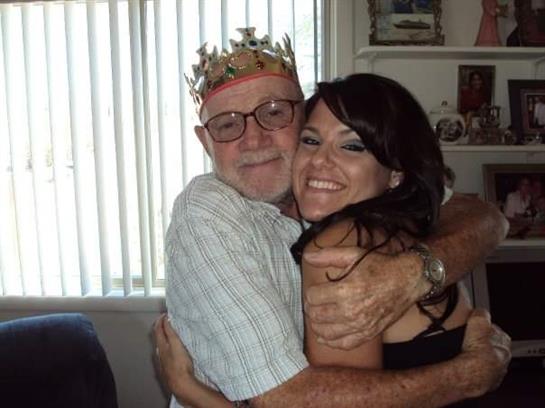 An elderly man wearing a crown shares a warm hug with a young woman in a bright room.