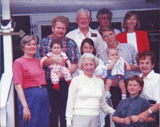 Family members including children and infants gather outside a house on a sunny day.